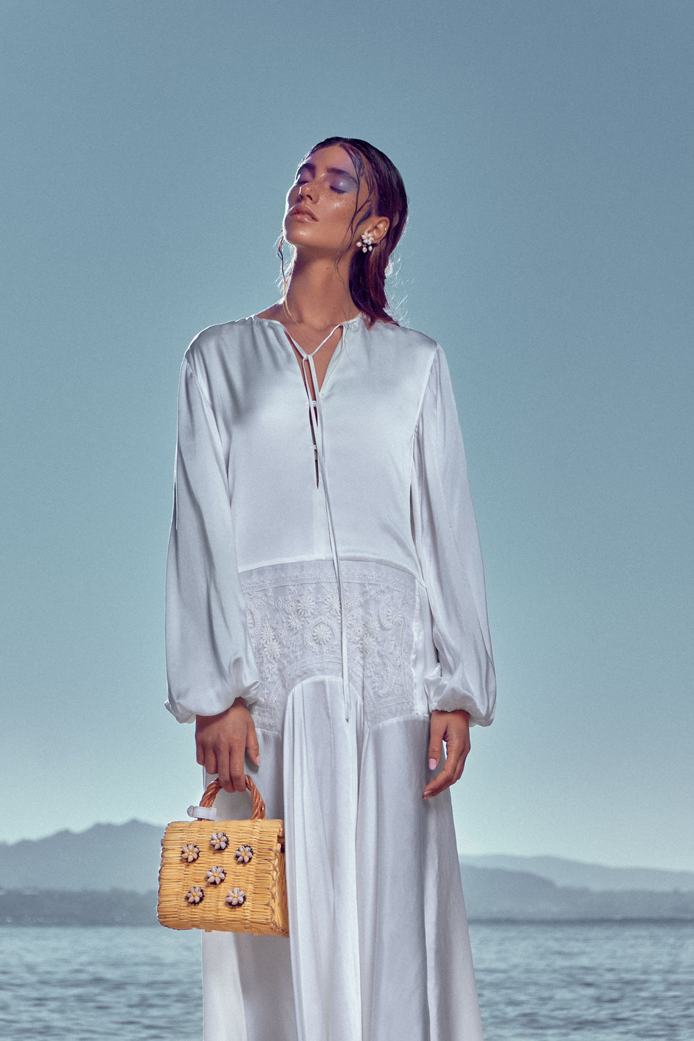 Shella Mini White basket bag by Heimat Atlantica, handwoven in Portugal from natural reed and finished in Spain with an Atlantic shell details. With model and Atlantics see behind. 